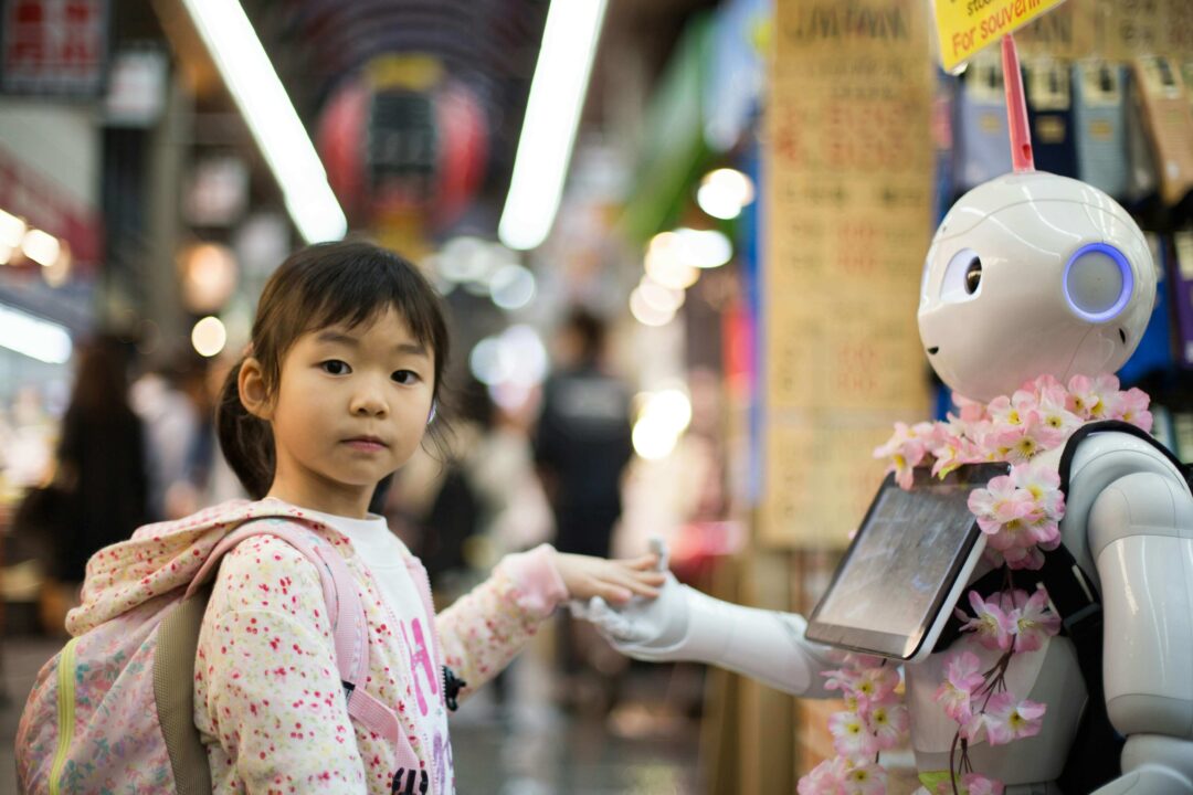 A little girl holding a vaguely humanoid robot's hand, blurred background (could be a shopping mall)