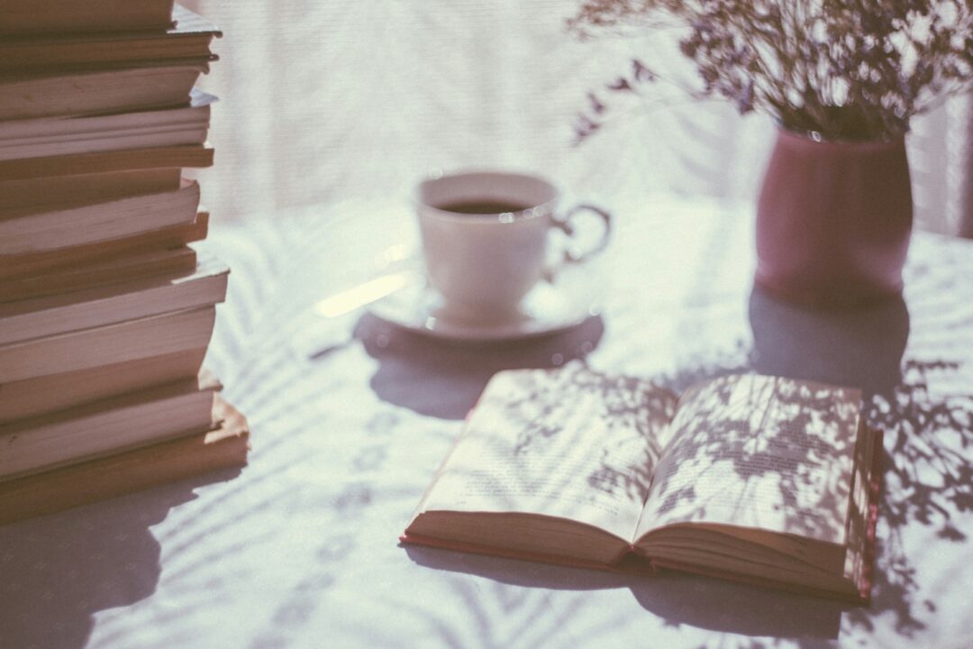 a generic stock photo of an open book, a stack of books (left) and a white ceramic coffee cup and vase of flowers in the background, all on a round table with white table cloth.